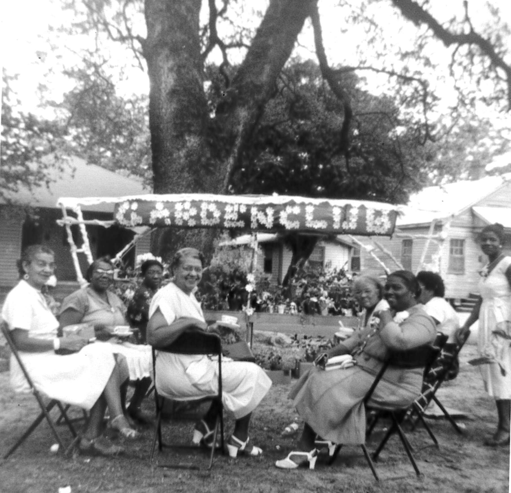 Eight black women smile towards the camera. They are seated in a semi-circle, dressed up and holding teacups. In front of them is a banner that says Garden Club.